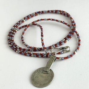Pode incluir: Um colar com um pingente de prata e uma corrente de contas. A corrente apresenta contas vermelhas, brancas e azuis. O pingente &eacute; uma moeda oval de prata com um design intrincado e um fecho.