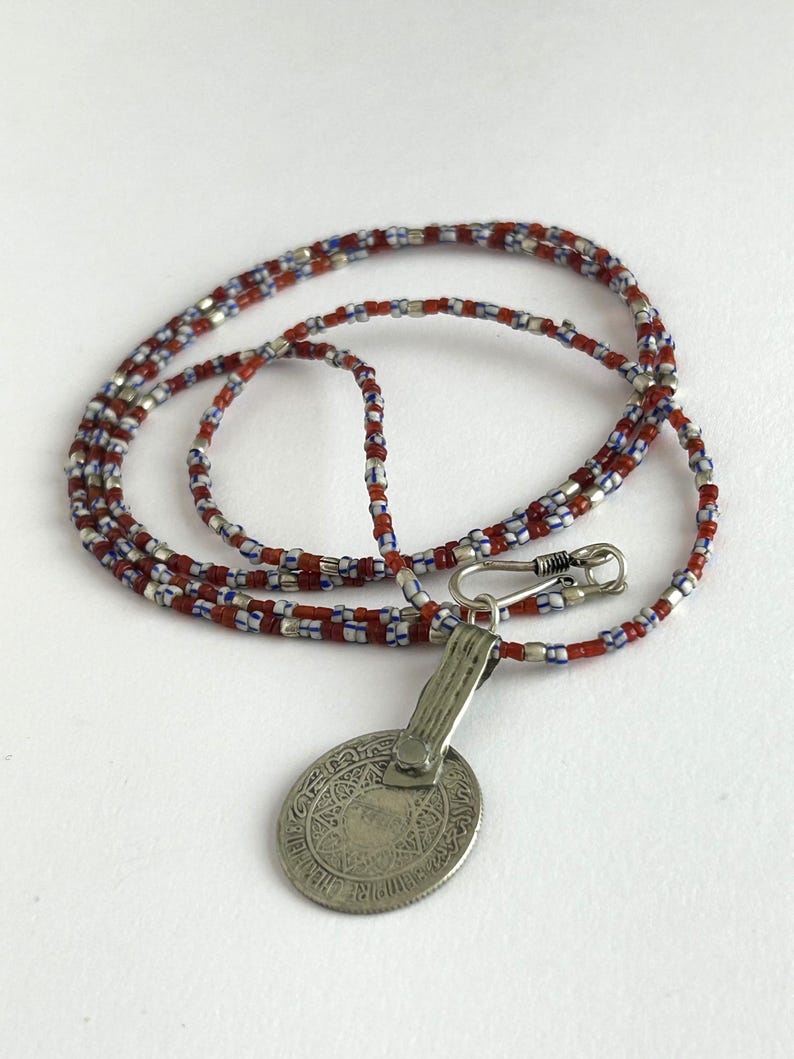 Pode incluir: Um colar com um pingente de prata e uma corrente de contas. A corrente apresenta contas vermelhas, brancas e azuis. O pingente &eacute; uma moeda oval de prata com um design intrincado e um fecho.