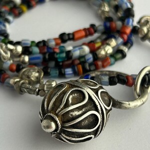 May include: A close-up of a necklace with a silver clasp and a large, ornate silver bead. The necklace features strands of colorful glass beads in various shapes and sizes, including red, blue, and black.