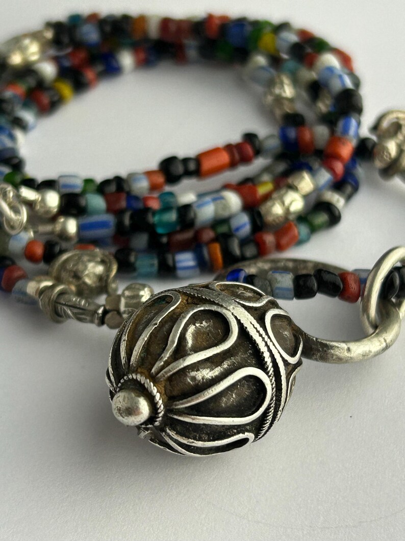 May include: A close-up of a necklace with a silver clasp and a large, ornate silver bead. The necklace features strands of colorful glass beads in various shapes and sizes, including red, blue, and black.