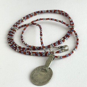 Pode incluir: Um colar com um pingente de prata e uma corrente de contas. A corrente apresenta contas vermelhas, brancas e azuis. O pingente &eacute; uma moeda oval de prata com um design intrincado e um fecho.