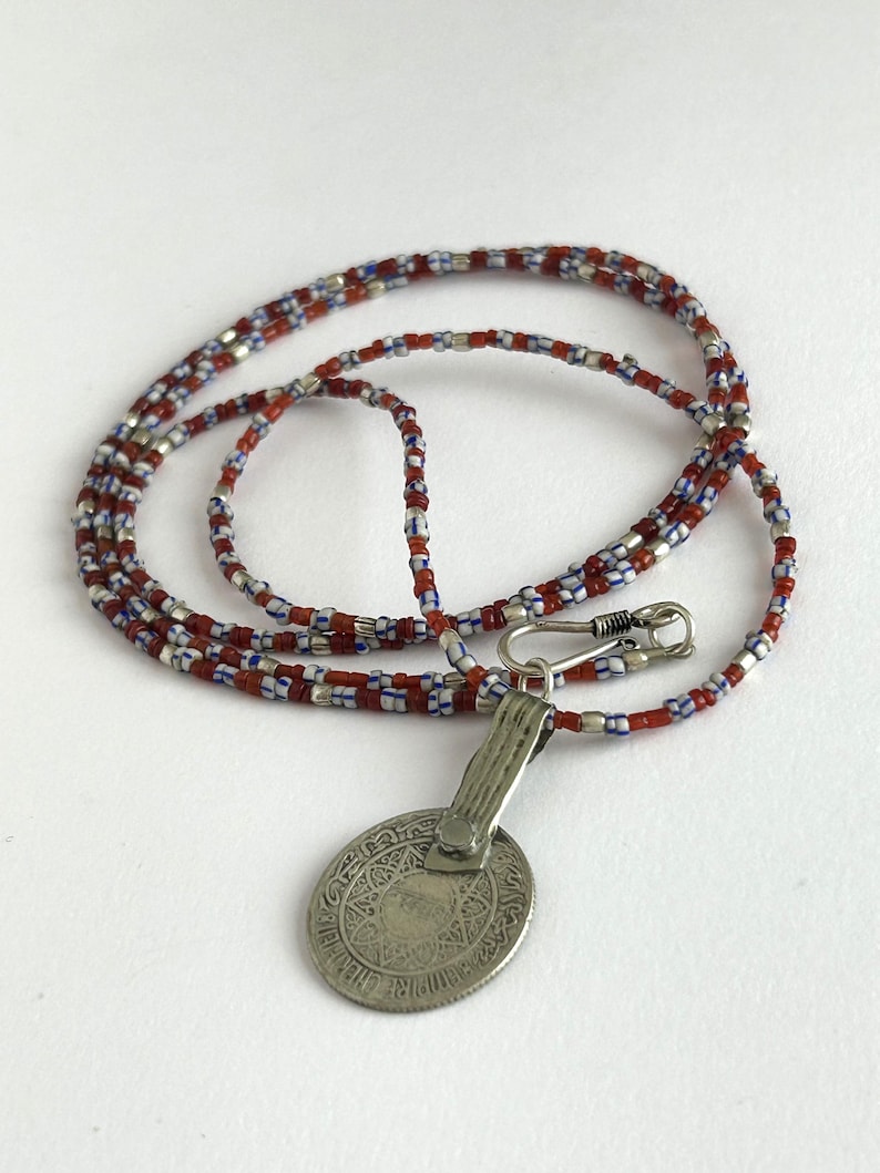 Pode incluir: Um colar com um pingente de prata e uma corrente de contas. A corrente apresenta contas vermelhas, brancas e azuis. O pingente &eacute; uma moeda oval de prata com um design intrincado e um fecho.
