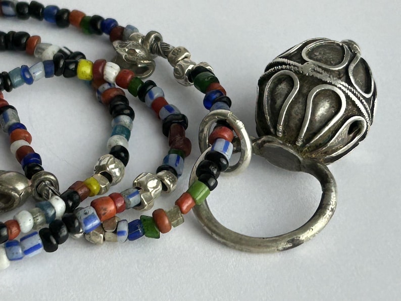 May include: Close-up of a necklace with colorful glass beads and silver accents. The necklace features a variety of small, round beads in red, blue, green, yellow, and black. A silver pendant with an ornate design is also visible.