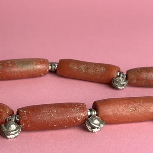 Pode incluir: Close-up de um colar com contas em forma de barril marrom-avermelhado. As contas s&atilde;o conectadas por elos de metal prateado com pingentes decorativos em forma de sino. Fundo rosa liso.