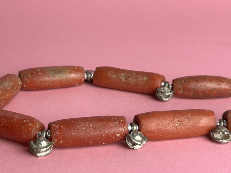 Pode incluir: Close-up de um colar com contas em forma de barril marrom-avermelhado. As contas s&atilde;o conectadas por elos de metal prateado com pingentes decorativos em forma de sino. Fundo rosa liso.