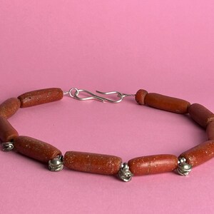 Pode incluir: Pulseira artesanal com contas cil&iacute;ndricas castanho-avermelhadas. As contas s&atilde;o unidas por pequenos detalhes met&aacute;licos prateados e um fecho prateado. A pulseira &eacute; exibida sobre um fundo rosa liso. As contas medem aproximadamente 1 cm de comprimento.