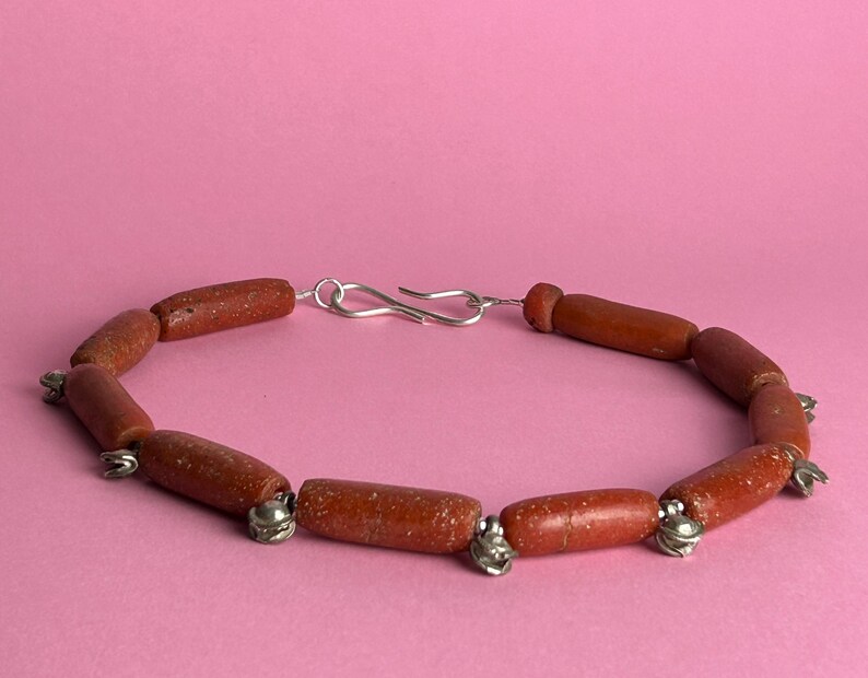 Pode incluir: Pulseira artesanal com contas cil&iacute;ndricas castanho-avermelhadas. As contas s&atilde;o unidas por pequenos detalhes met&aacute;licos prateados e um fecho prateado. A pulseira &eacute; exibida sobre um fundo rosa liso. As contas medem aproximadamente 1 cm de comprimento.