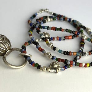 May include: A silver pendant with an ornate, textured design, connected to a silver ring. The pendant is paired with a multi-strand necklace composed of small, colorful beads in various hues, including red, blue, green, and black. The necklace is strung with silver accents.