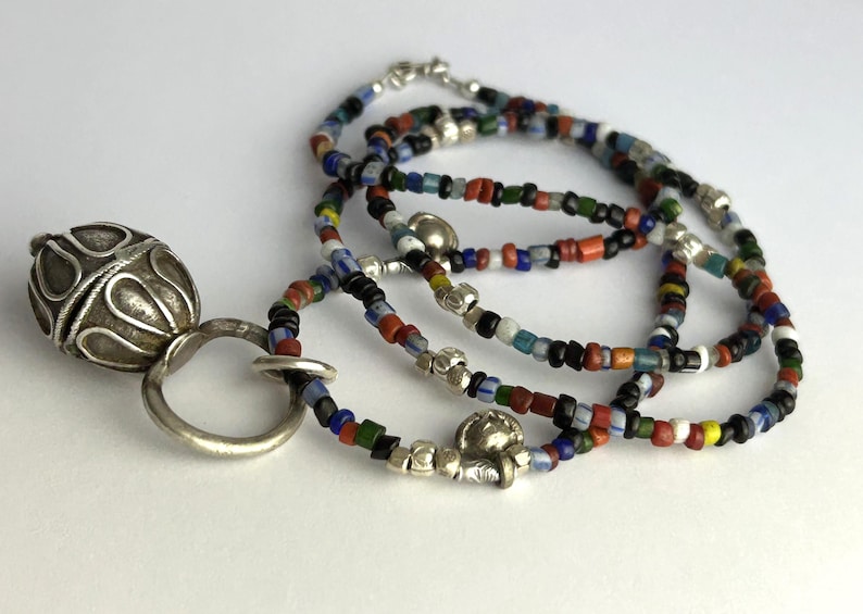 May include: A silver pendant with an ornate, textured design, connected to a silver ring. The pendant is paired with a multi-strand necklace composed of small, colorful beads in various hues, including red, blue, green, and black. The necklace is strung with silver accents.