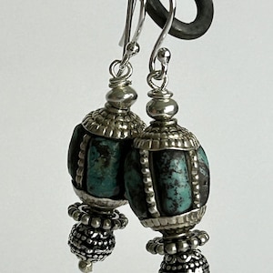 May include: A pair of silver earrings with turquoise-colored beads. The earrings have a detailed design with a combination of silver and turquoise elements. The earrings are suspended from silver hooks.