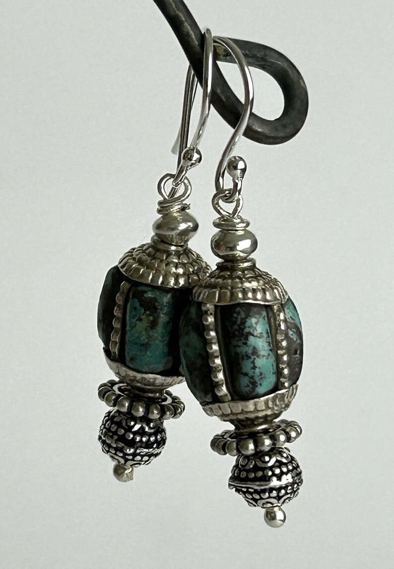 May include: A pair of silver earrings with turquoise-colored beads. The earrings have a detailed design with a combination of silver and turquoise elements. The earrings are suspended from silver hooks.