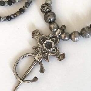 May include: A silver-toned brooch with a floral design and a pin, paired with a beaded necklace featuring gray and black beads. The brooch has a central flower shape with detailed petals and a circular center. The necklace has multiple strands.