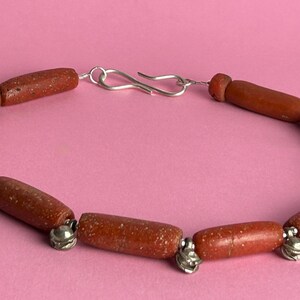 Pode incluir: Uma pulseira feita com contas cil&iacute;ndricas castanho-avermelhadas. As contas s&atilde;o unidas com pequenos detalhes met&aacute;licos prateados e um fecho prateado. A pulseira &eacute; exibida sobre um fundo rosa.