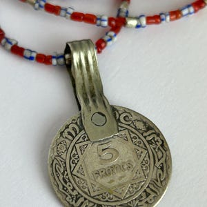 Pode incluir: Colar com pingente de moeda de prata e corrente de contas. A moeda apresenta o n&uacute;mero "5" e a palavra "FRANCS" dentro de um desenho em forma de estrela. A corrente do colar &eacute; feita de contas vermelhas, brancas e azuis.