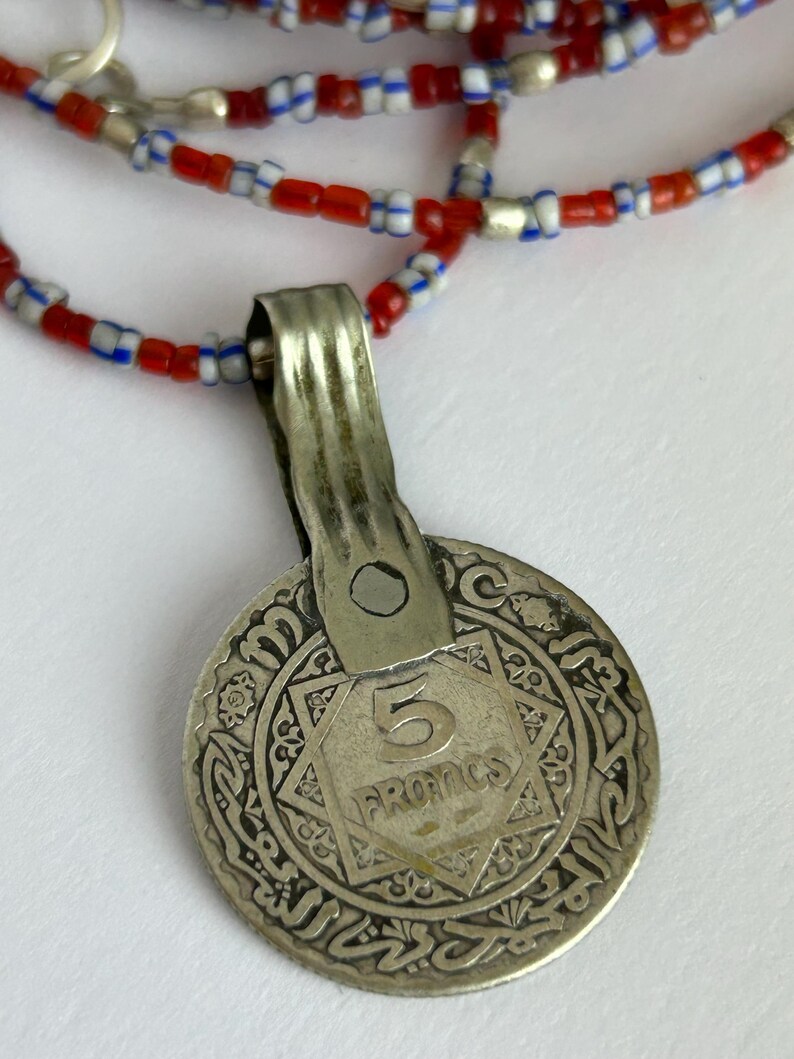 Pode incluir: Colar com pingente de moeda de prata e corrente de contas. A moeda apresenta o n&uacute;mero "5" e a palavra "FRANCS" dentro de um desenho em forma de estrela. A corrente do colar &eacute; feita de contas vermelhas, brancas e azuis.