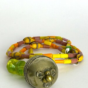 May include: A silver-colored metal bottle stopper with a decorative floral design and a green glass stopper. A necklace with yellow, orange, and pink beads is also in the image. The beads are strung together with a yellow cord.