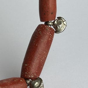 Pode incluir: Close-up de um colar com contas cil&iacute;ndricas castanho-avermelhadas. As contas s&atilde;o conectadas por fechos prateados ornamentados. O colar tem uma est&eacute;tica r&uacute;stica e terrosa.