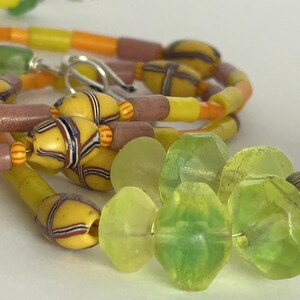 May include: Close-up of a colorful necklace featuring yellow, orange, and pink beads. The necklace has a variety of shapes and sizes, including cylindrical and patterned beads. Green, faceted beads are also visible, along with a silver clasp.