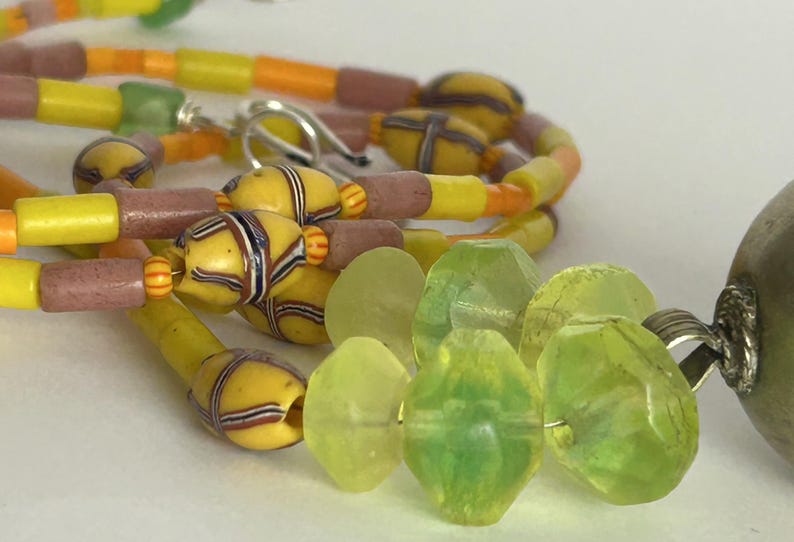 May include: Close-up of a colorful necklace featuring yellow, orange, and pink beads. The necklace has a variety of shapes and sizes, including cylindrical and patterned beads. Green, faceted beads are also visible, along with a silver clasp.