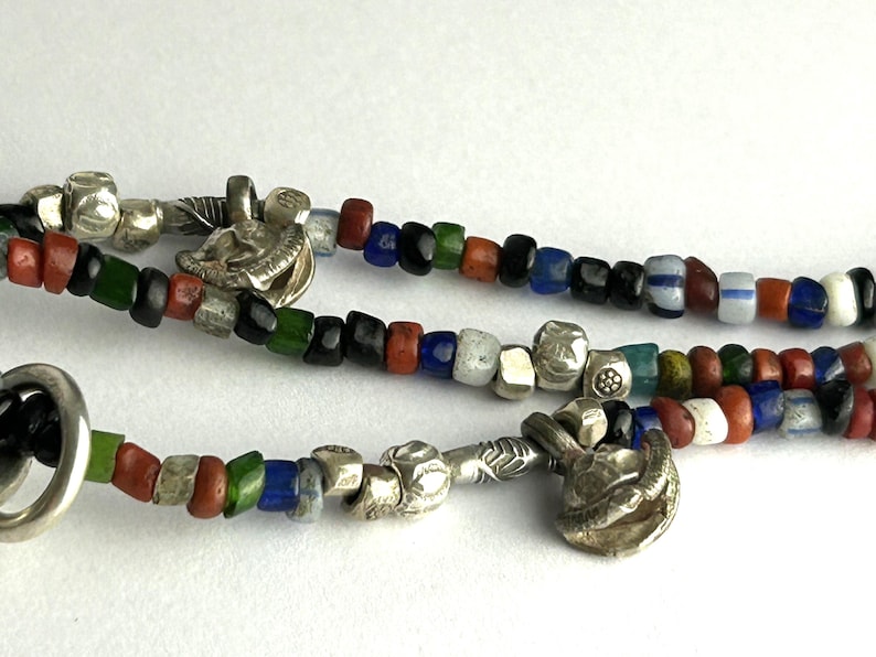 May include: Close-up of three strands of colorful beaded necklaces. The beads are in various colors, including red, green, blue, and white. Silver-toned metal charms and clasps are interspersed throughout the strands.