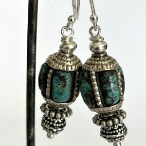 May include: A pair of silver earrings with turquoise-colored beads. The earrings have a detailed design with a silver cap and a beaded bottom. The turquoise beads have dark veining. The earrings are hanging from a metal stand.