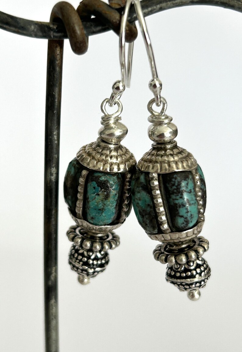 May include: A pair of silver earrings with turquoise-colored beads. The earrings have a detailed design with a silver cap and a beaded bottom. The turquoise beads have dark veining. The earrings are hanging from a metal stand.