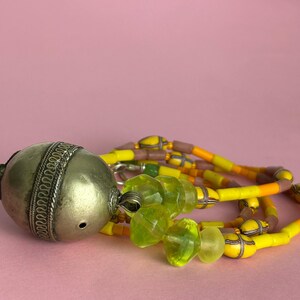 May include: An antique silver-toned metal pendant with intricate detailing, connected to a necklace of yellow, orange, and brown beads. The necklace also features translucent green glass beads. The pendant is resting on a pink surface.