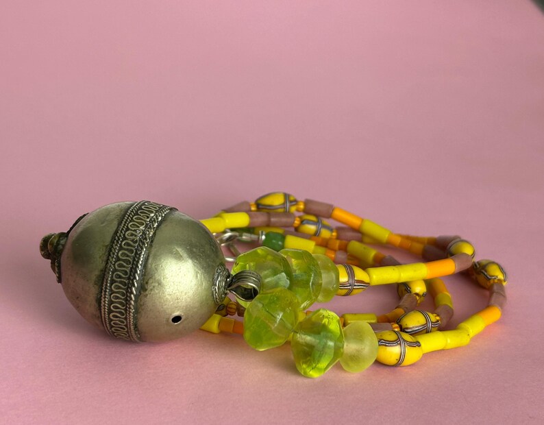 May include: An antique silver-toned metal pendant with intricate detailing, connected to a necklace of yellow, orange, and brown beads. The necklace also features translucent green glass beads. The pendant is resting on a pink surface.