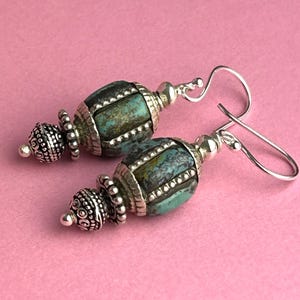 May include: A pair of turquoise and silver earrings. The earrings feature a turquoise-colored, barrel-shaped bead with silver accents. The silver accents include decorative caps and a hook-style ear wire. The background is a solid pink color.