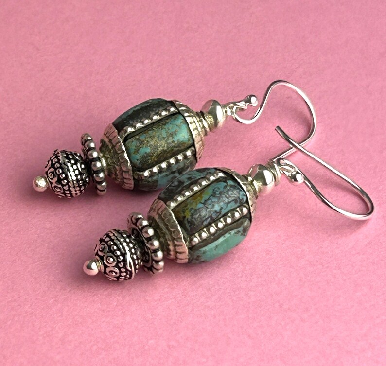 May include: A pair of turquoise and silver earrings. The earrings feature a turquoise-colored, barrel-shaped bead with silver accents. The silver accents include decorative caps and a hook-style ear wire. The background is a solid pink color.