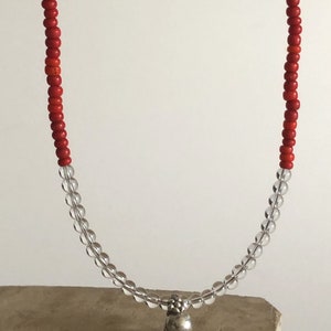 May include: A red and clear beaded necklace with a silver charm. The necklace is on a brown surface.