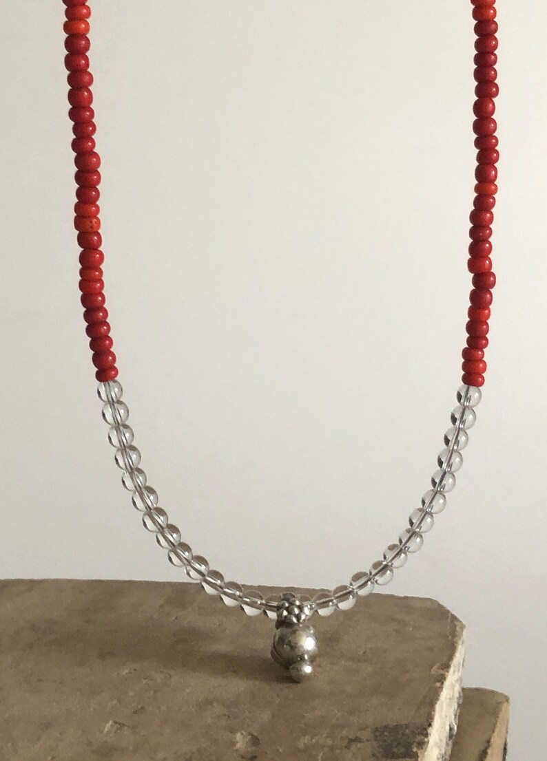 May include: A red and clear beaded necklace with a silver charm. The necklace is on a brown surface.