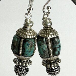 May include: A pair of silver earrings with turquoise and black stone beads. The earrings have a decorative silver cap and a detailed silver bead at the bottom. The earrings hang from a silver hook.