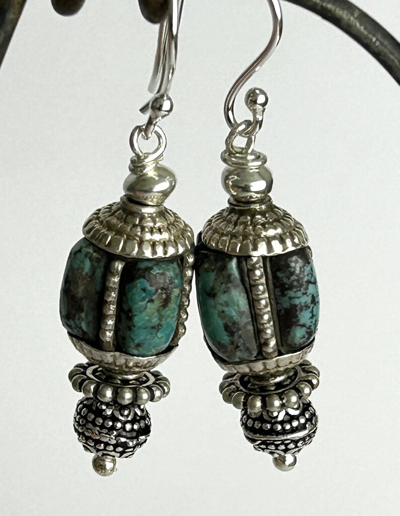 May include: A pair of silver earrings with turquoise and black stone beads. The earrings have a decorative silver cap and a detailed silver bead at the bottom. The earrings hang from a silver hook.