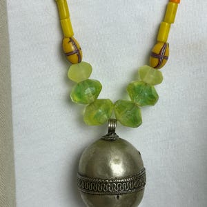 May include: A handcrafted necklace featuring a silver pendant with a decorative band and a beaded strand. The necklace has yellow, orange, and green beads. The pendant is a silver oval shape.