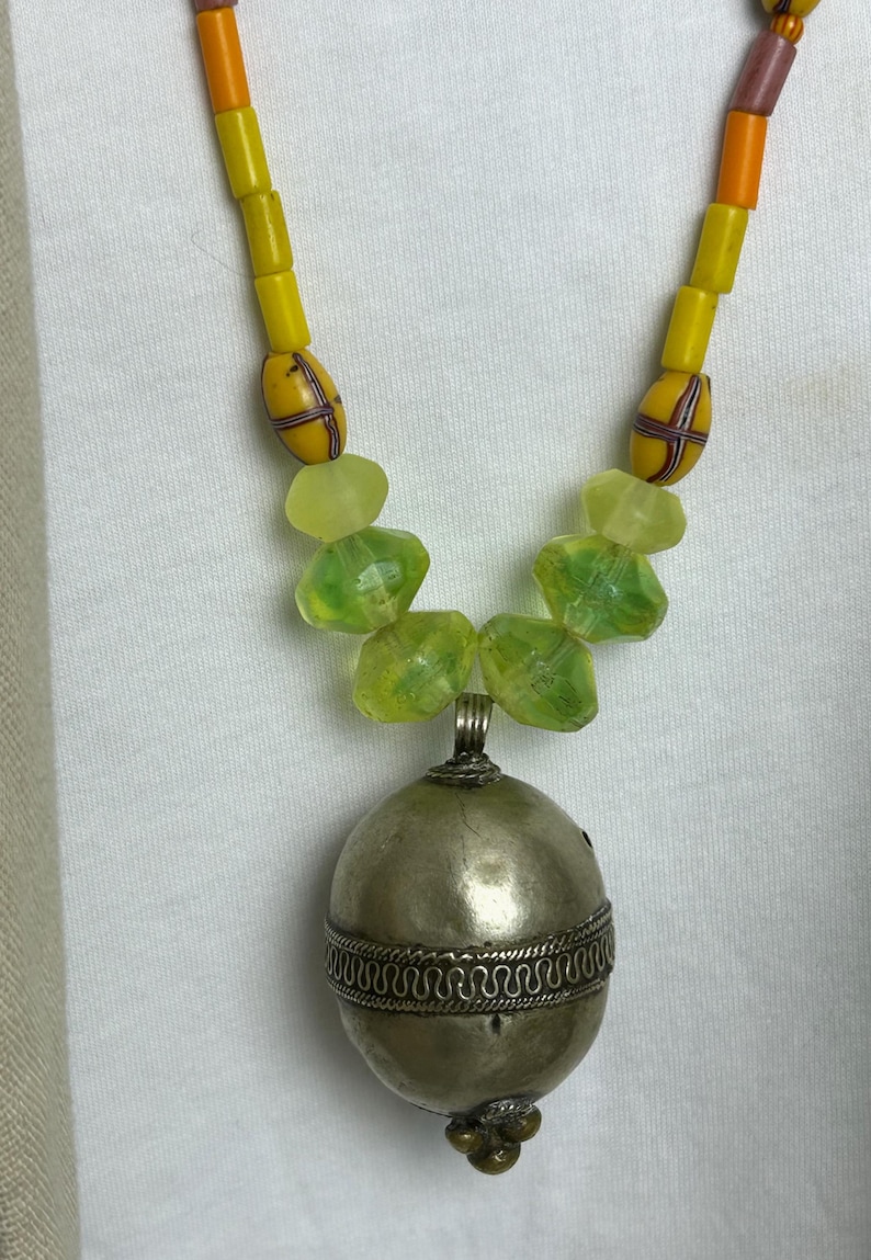May include: A handcrafted necklace featuring a silver pendant with a decorative band and a beaded strand. The necklace has yellow, orange, and green beads. The pendant is a silver oval shape.