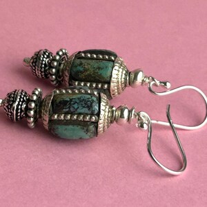 May include: A pair of handcrafted earrings featuring turquoise-colored stones and ornate silver-toned metalwork. The earrings have a barrel-shaped design with decorative caps and hook closures, set against a pink background.