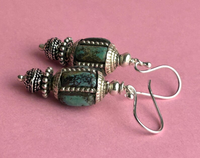 May include: A pair of handcrafted earrings featuring turquoise-colored stones and ornate silver-toned metalwork. The earrings have a barrel-shaped design with decorative caps and hook closures, set against a pink background.