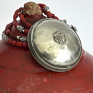 May include: A round silver pendant with a central floral motif and a beaded edge, attached to a multi-strand necklace featuring small red cylindrical beads interspersed with decorative silver spacers, creating a layered and textured appearance.