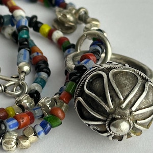 May include: A close-up of a colorful beaded necklace with a silver pendant. The necklace features a variety of small, multi-colored beads in red, blue, green, yellow, and black. The silver pendant is a round, ornate design.