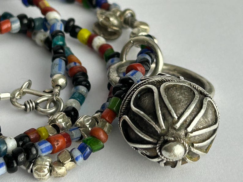 May include: A close-up of a colorful beaded necklace with a silver pendant. The necklace features a variety of small, multi-colored beads in red, blue, green, yellow, and black. The silver pendant is a round, ornate design.