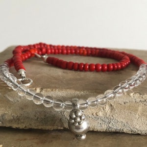 May include: A layered necklace with clear glass beads and a silver pendant. The top layer is made of red beads.
