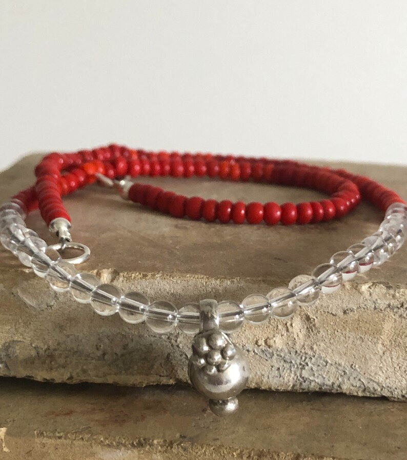May include: A layered necklace with clear glass beads and a silver pendant. The top layer is made of red beads.