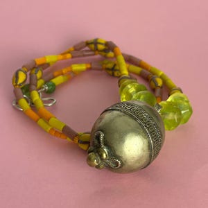 May include: A necklace featuring a large, silver-colored metal pendant with a decorative design. The necklace has strands of yellow, orange, and brown beads, with a few green beads. The background is a solid pink color.