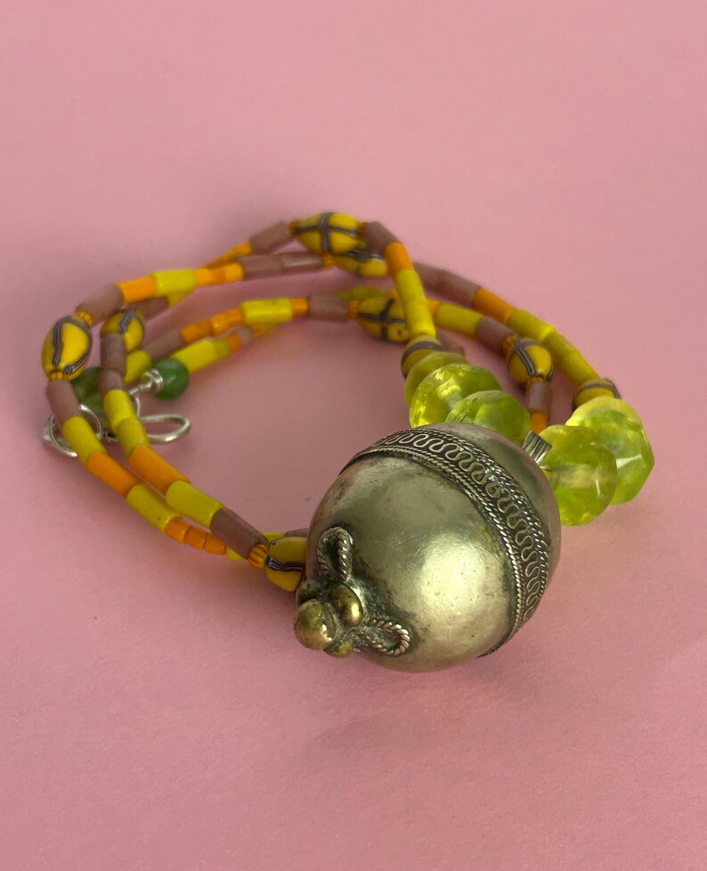May include: A necklace featuring a large, silver-colored metal pendant with a decorative design. The necklace has strands of yellow, orange, and brown beads, with a few green beads. The background is a solid pink color.