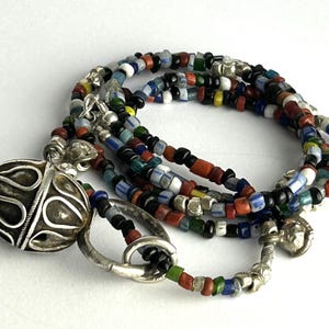 May include: A multi-strand necklace featuring colorful glass beads in red, black, blue, green, and yellow. The necklace has a silver-toned filigree pendant and clasp. The beads are small and cylindrical, creating a layered effect.