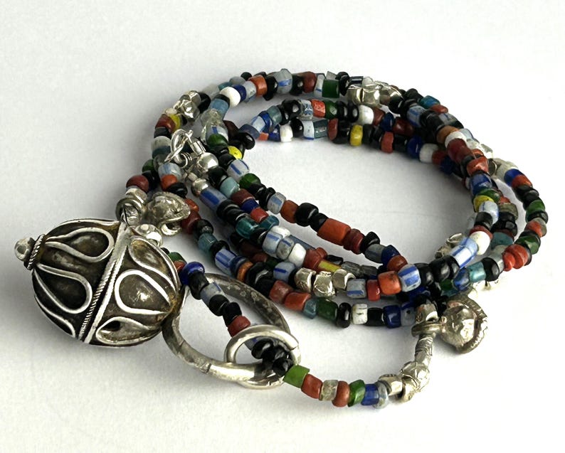 May include: A multi-strand necklace featuring colorful glass beads in red, black, blue, green, and yellow. The necklace has a silver-toned filigree pendant and clasp. The beads are small and cylindrical, creating a layered effect.