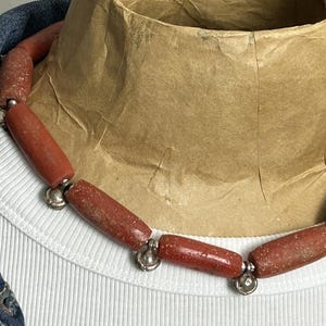 Pode incluir: Um colar com contas cil&iacute;ndricas castanho-avermelhadas com uma textura salpicada. As contas s&atilde;o unidas com detalhes met&aacute;licos prateados. O colar &eacute; exibido em uma camisa branca canelada e uma jaqueta jeans.