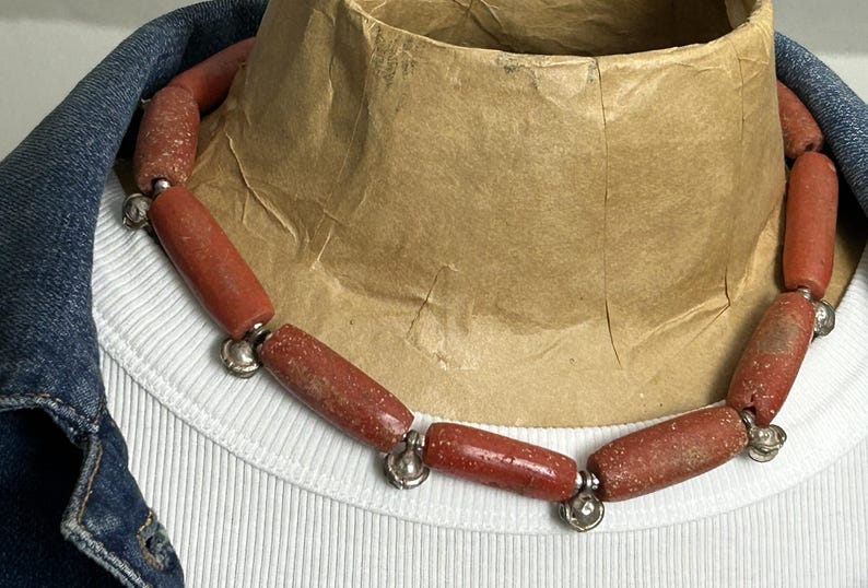 Pode incluir: Um colar com contas cil&iacute;ndricas castanho-avermelhadas com uma textura salpicada. As contas s&atilde;o unidas com detalhes met&aacute;licos prateados. O colar &eacute; exibido em uma camisa branca canelada e uma jaqueta jeans.