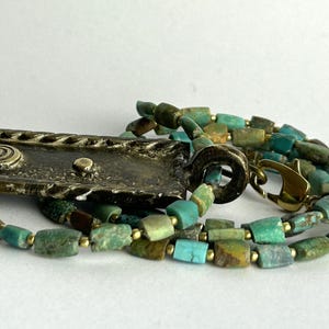May include: A necklace featuring a rectangular, textured bronze-toned pendant with a spiral design, attached to a strand of small, irregular turquoise and green stone beads interspersed with tiny gold-toned metal spacers and a lobster clasp.